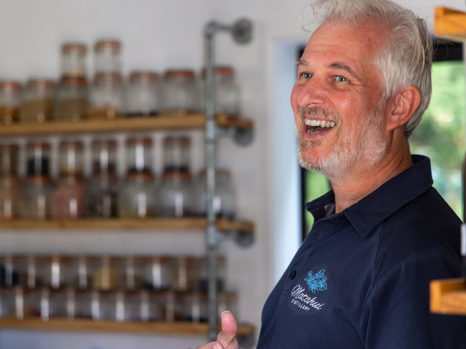 Paul mid-conversation at Matahui Distillery, laughing, with shelves of botanical infusion jars behind him.
